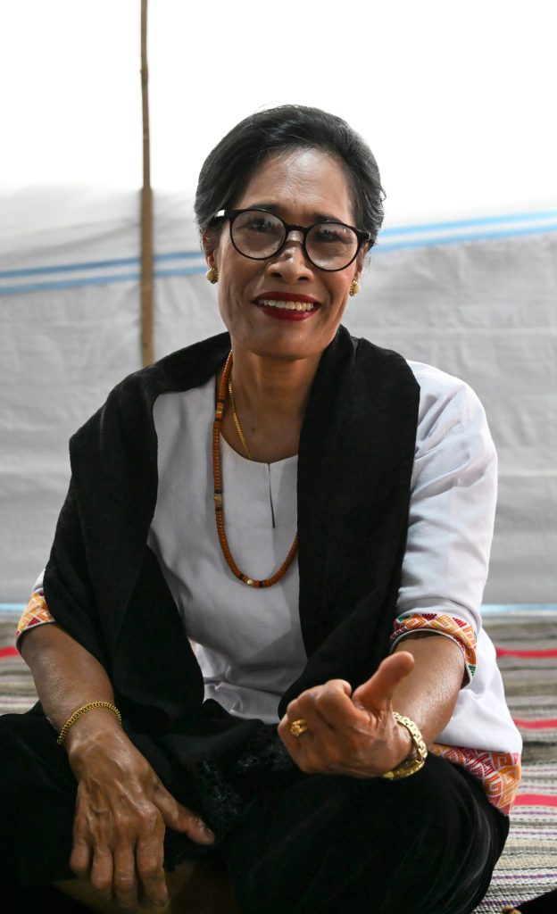 A portrait of Berta’s daughter sitting inside a shaded traditional Torajan house (Tongkonan) with a warm smile and an "OK" gesture, wearing glasses and a traditional white and black outfit.