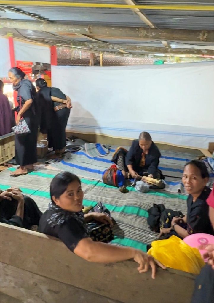 A frame showing the back-view of several people sitting on woven mats inside a shaded traditional Tongkonan house structure during a Tana Toraja funeral ceremony.