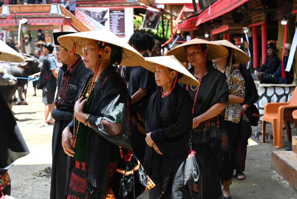 A solemn line of three Torajan women in traditional black robes and wide 'caping' woven hats walks in procession during a funeral, their faces showing profound sorrow.