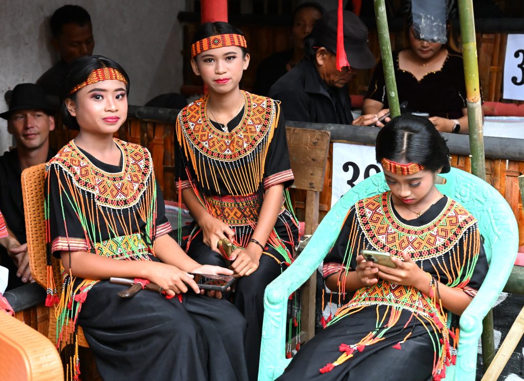 A group of three young Torajan girls dressed in detailed traditional black and red beaded collars and headbands. One girl on the right is looking down at a smartphone, while others sit on simple plastic chairs during a funeral ceremony in Sulawesi.