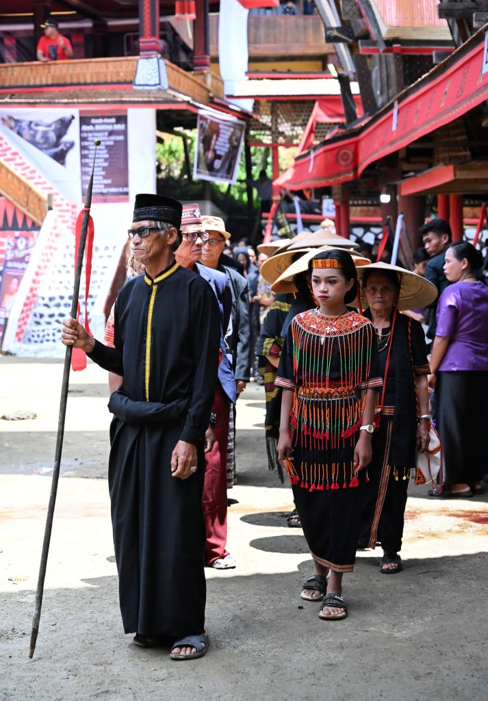 An elderly Torajan man in a black robe and peci cap leads a funeral procession with a staff, followed by a young woman in beaded traditional dress and older women in conical caping hats.
