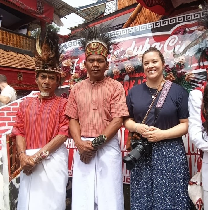 A female traveler standing and smiling between two Torajan men in traditional ceremonial dress, featuring red striped tunics, white sarongs, and tall feather headpieces during a Rambu Solo funeral.