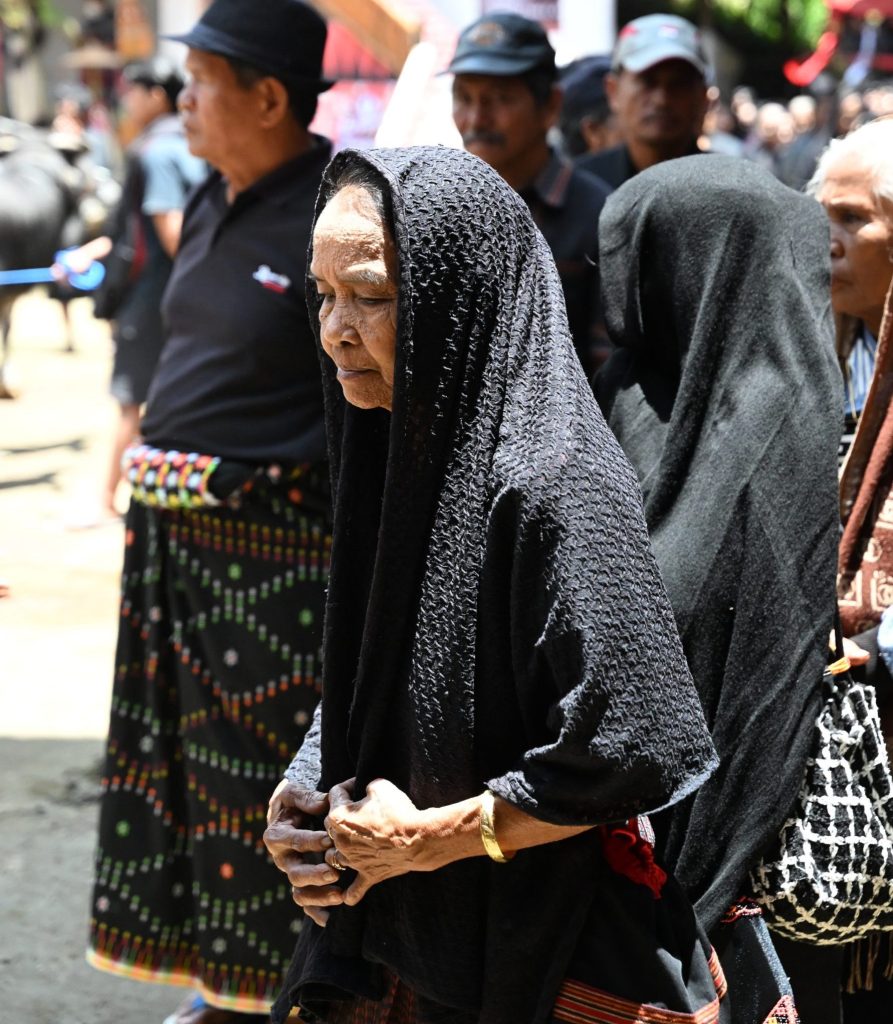 A close-up portrait of an elderly Torajan woman in a black patterned veil, her face a landscape of sorrow and memory as she walks in a funeral procession. Her hands are clasped at her waist.