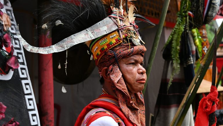 A horizontal portrait of a Torajan ceremonial leader with a determined expression. He wears an elaborate feathered headpiece, a red ceremonial wrap, and stands among traditional warriors holding spears, against a backdrop of traditional curved-roof houses.