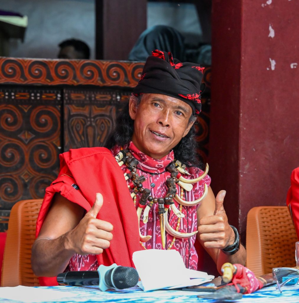 A middle-aged Torajan man acting as the Master of Ceremonies at a Tana Toraja funeral. He is wearing a traditional red patterned shirt and a distinctive tall feather headpiece, holding a microphone and speaking with a focused, commanding expression.
