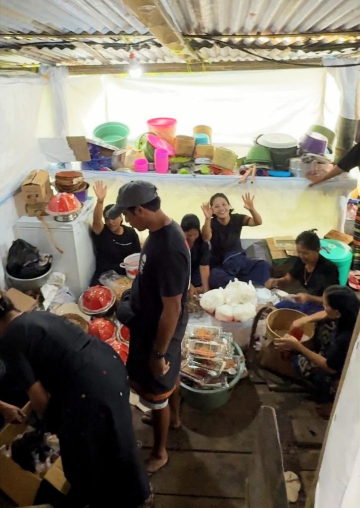 A high-angle frame from a video shows the inside of a bustling Torajan Tongkonan kitchen. A group of men and women sit on the floor preparing food, surrounded by kitchen supplies, waving and smiling toward the camera. A metal corrugated roof is overhead.