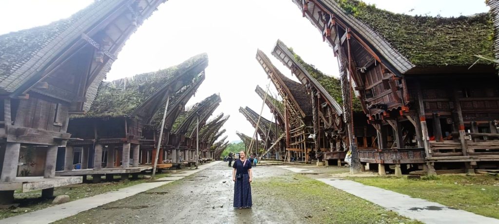 The author standing in the center of a dirt path lined on both sides by massive, traditional Torajan Tongkonan houses with their signature boat-shaped roofs reaching toward the sky.
