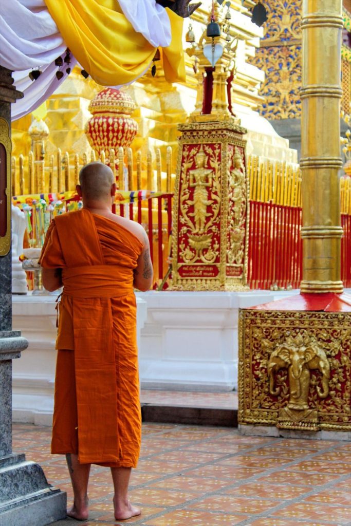 A Buddhist monk in vibrant orange robes seen from behind, standing barefoot on patterned tile and looking toward the intricate golden chedi and red ceremonial pillars at Wat Phra That Doi Suthep.