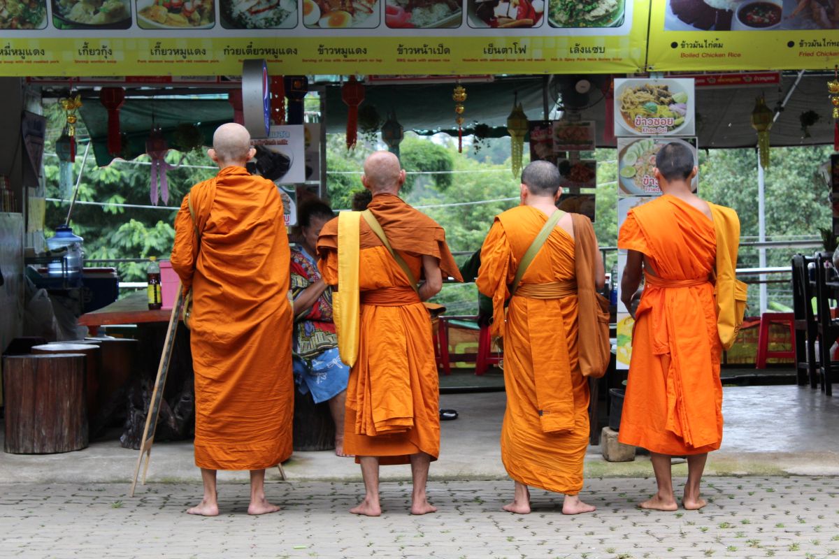 Four Buddhist monks in orange robes seen from behind, standing in a line at a local food stall with Thai menu signs, during their morning alms walk in the community.