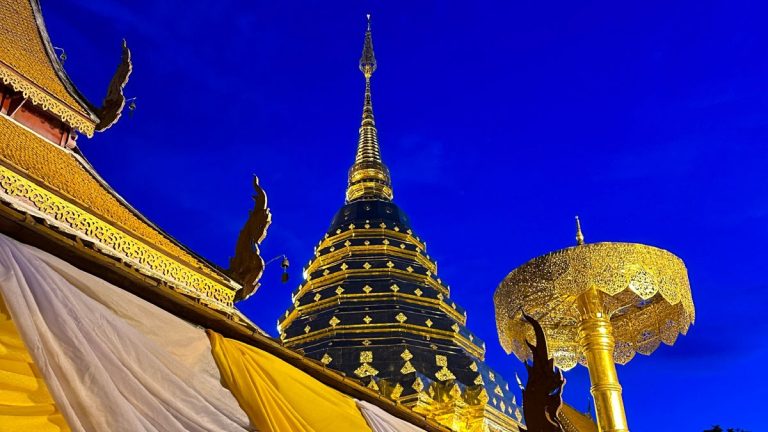 The iconic golden Lanna-style chedi (stupa) of Wat Phra That Doi Suthep illuminated against a deep navy blue sky, with intricate gold carvings and yellow ceremonial fabric in the foreground.