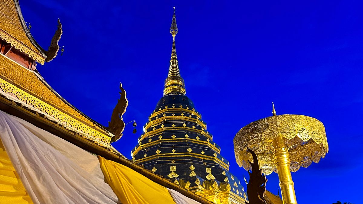 The iconic golden Lanna-style chedi (stupa) of Wat Phra That Doi Suthep illuminated against a deep navy blue sky, with intricate gold carvings and yellow ceremonial fabric in the foreground.