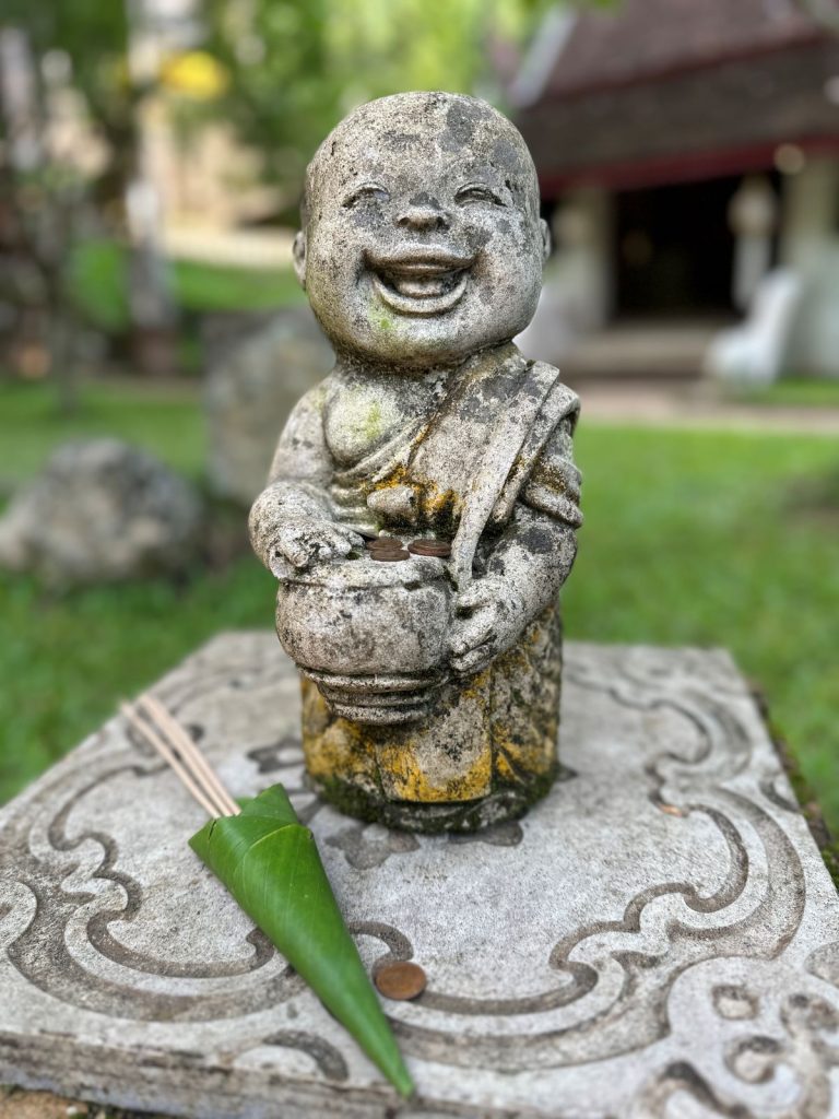 A small, joyful stone statue of a laughing novice monk holding an alms bowl with coins, located in the jungle gardens of Wat Pha Lat in Chiang Mai.