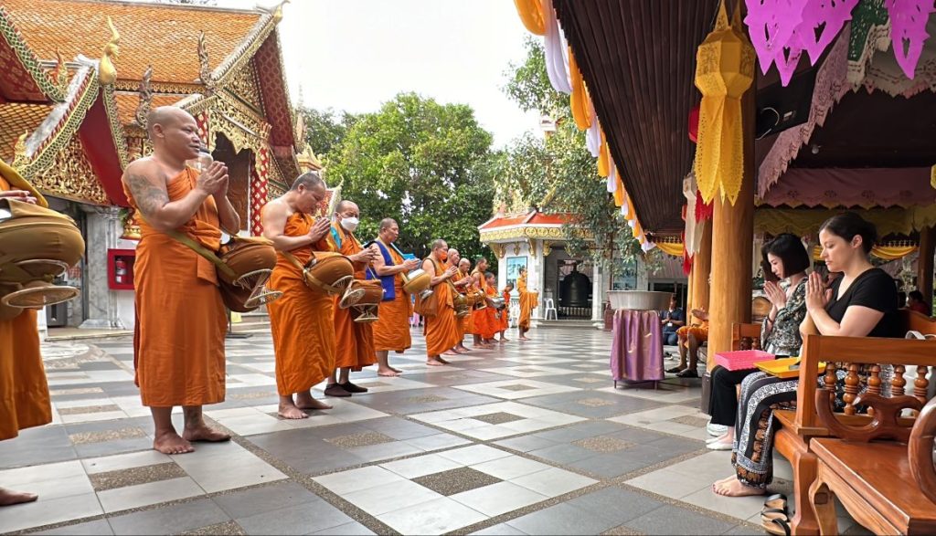 A line of Buddhist monks in saffron robes standing with alms bowls, facing a row of seated visitors including the author in a black shirt, all with hands pressed together in a respectful prayer gesture.