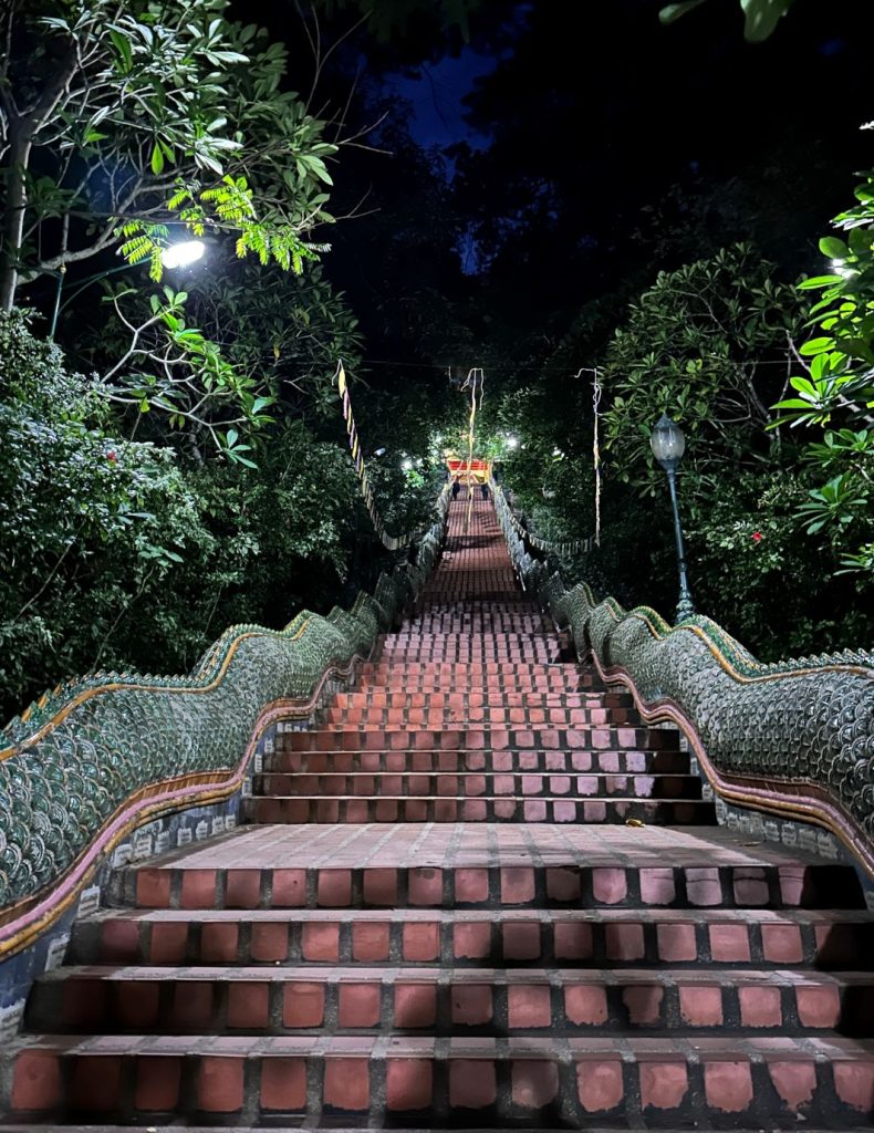 A ground-level view looking up the long, steep 306-step staircase of Wat Phra That Doi Suthep at a pre-dawn hour, flanked by ornate green Naga serpent balustrades and lit by glowing streetlamps under a dark sky.