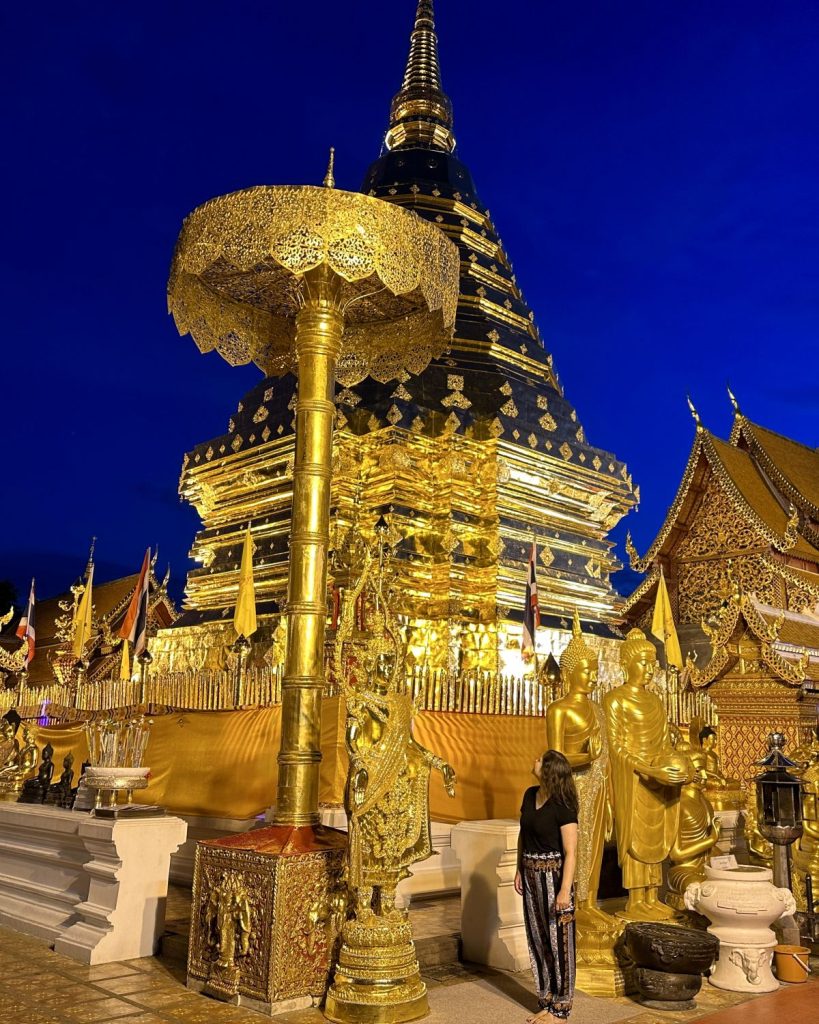 A full-body portrait of a female traveler, looking back to the temple, dressed in patterned pants and a black shirt. She is at Wat Phra That Doi Suthep temple at blue hour, positioned next to ornate golden parasols and standing statues, with the massive illuminated golden chedi (stupa) glowing under a deep blue sky.